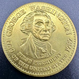 May include: A gold-colored coin with a portrait of George Washington, the first president of the United States. The coin reads "George Washington 1789 First President of the United States 1797".