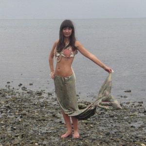 Puede incluir: Una mujer con un sujetador de concha marina y una falda de lentejuelas con cola de sirena. Est&aacute; de pie en una playa rocosa con el oc&eacute;ano de fondo.