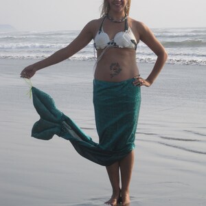 Puede incluir: Una mujer con un top de bikini blanco y negro de conchas marinas y una cola de sirena turquesa en una playa.