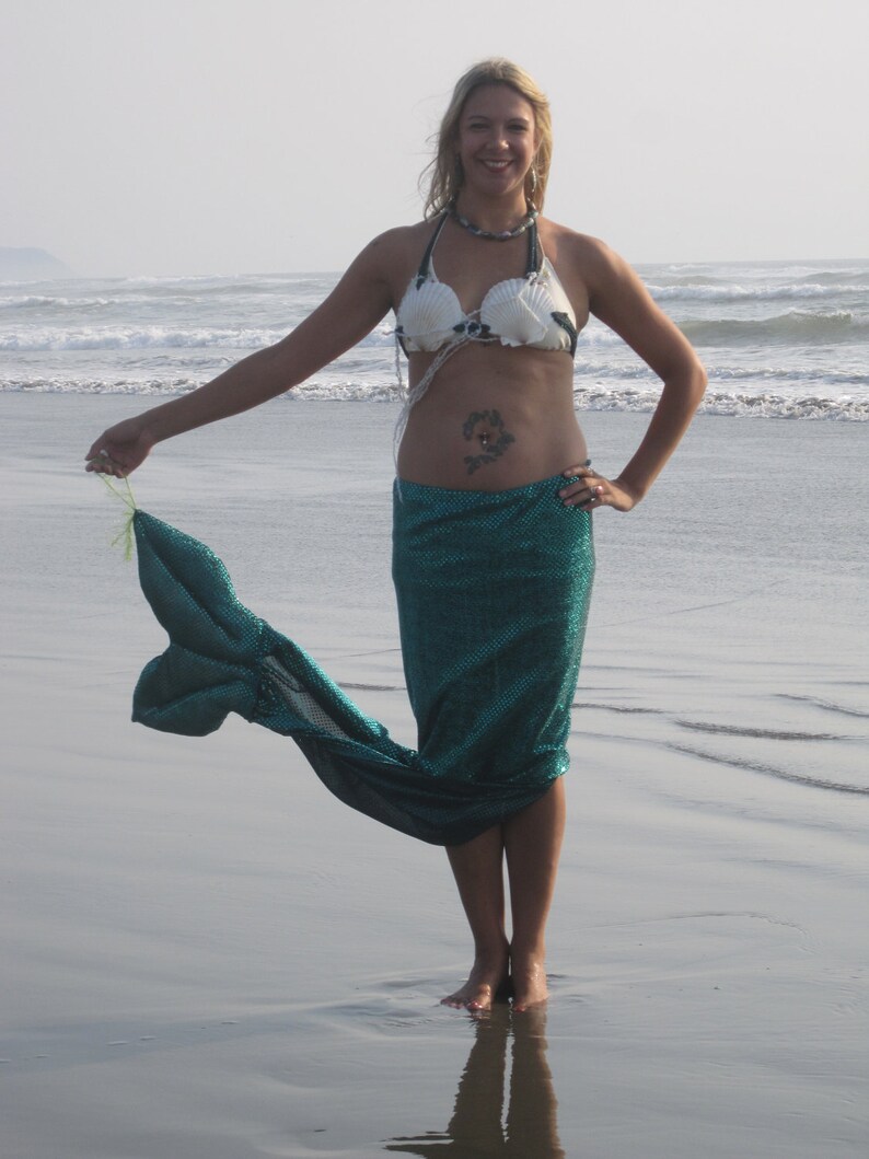 Puede incluir: Una mujer con un top de bikini blanco y negro de conchas marinas y una cola de sirena turquesa en una playa.