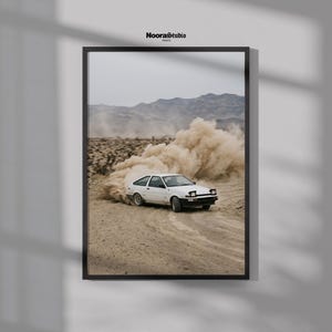 May include: A framed wall art print featuring a white hatchback car drifting on a dirt road, kicking up a large cloud of dust. The scene is set in a desert landscape with mountains in the background. The text NooraStudio is printed at the top.