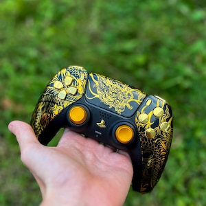 May include: A black and gold PlayStation 5 controller with a dragon design. The controller has yellow buttons and a gold PlayStation logo.