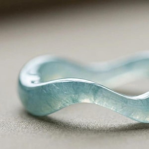 May include: A translucent, light blue ring with a smooth, wavy, undulating band design. The material has a glossy, polished finish that catches the light, revealing subtle internal textures and variations within the pale blue, glass-like structure.