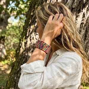 Puede incluir: Una pulsera de cuentas multicolores con cuentas rojas, verdes, amarillas y azules. La pulsera se lleva en la muñeca, con el brazo y la mano de una persona visibles. La persona lleva una camisa blanca y está cerca de un árbol.