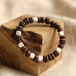 Puede incluir: Una pulsera circular compuesta por granos de café tostados de color marrón oscuro intercalados con pequeñas cuentas cuadradas blancas. Cada cuenta blanca presenta una letra dorada, formando la secuencia J, A, J, S, E, D y S.