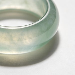 May include: A close-up of a translucent jade ring. The ring is a pale green color with a smooth, polished surface. The light passes through the jade, creating a soft glow. The ring is a solid band, with a rounded shape.