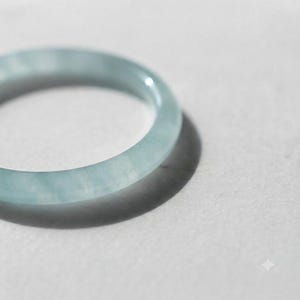 Puede incluir: Un anillo circular liso elaborado con piedra de color azul claro pálido y translúcido. La banda presenta un acabado pulido con sutiles variaciones de color internas similares a nubes, creando una apariencia suave y consistente en toda la forma.