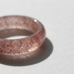 Puede incluir: Un anillo circular hecho de un material translúcido de color rosa pálido, incrustado con numerosas motas pequeñas de color marrón rojizo oscuro en toda su estructura. La banda tiene una superficie lisa y redondeada con un acabado brillante.