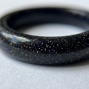 May include: A close-up of a dark blue ring with a speckled, glittery appearance. The ring is a solid band, likely made of stone or resin, and has a smooth, polished surface. The ring's colour is a deep, rich blue with gold flecks.