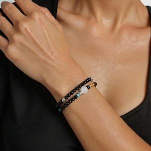 Puede incluir: Una pulsera de cuentas de doble hilo con pequeñas cuentas negras pulidas, acentuada con una cuenta blanca más grande, una cuenta turquesa, una cuenta naranja y espaciadores de metal plateado integrados en todo el diseño.