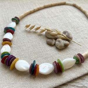 Puede incluir: Un collar artesanal con un cordón beige y una serie de cuentas blancas en forma de lágrima, intercaladas con detalles circulares coloridos en tonos rojos, verdes, amarillos y morados. El collar se muestra sobre una superficie de lino neutra.