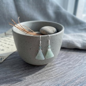 Puede incluir: Un par de pendientes colgantes con colgantes de piedra triangulares de color verde pálido suspendidos de ganchos plateados. Cada pendiente incluye una pequeña cuenta redonda verde a juego colocada sobre el componente de piedra triangular.