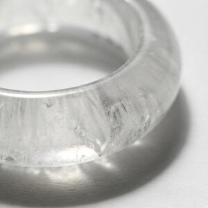 Puede incluir: Un anillo transparente e incoloro con una banda redondeada y lisa. El material es translúcido y presenta fracturas internas e inclusiones naturales que crean una apariencia cristalina y texturizada en toda la estructura circular.