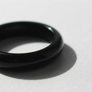 Puede incluir: Un anillo negro liso y sólido con un acabado pulido y brillante. La banda presenta un perfil circular redondeado con un grosor constante, reflejando la luz en su superficie curva para resaltar el color oscuro del material.