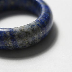 Puede incluir: Un anillo pulido elaborado en piedra lapislázuli de color azul profundo, que presenta una banda lisa y redondeada con inclusiones naturales visibles de motas de pirita gris y dorada distribuidas por la superficie del material de piedra.