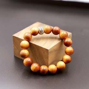 Puede incluir: Una pulsera de cuentas de madera con cuentas grandes y redondas en tonos marrones y naranjas. La pulsera presenta una cuenta de acento plateada con un diseño intrincado. Se muestra sobre un bloque de madera sobre un fondo oscuro.