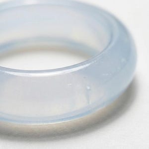 Puede incluir: Un anillo liso y translúcido con un tono azul hielo pálido. La banda presenta una superficie redondeada y pulida que capta la luz, creando sutiles reflejos a lo largo de sus bordes curvos. El material tiene una apariencia clara y vítrea.