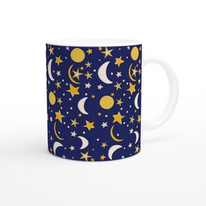 May include: White ceramic mug with a repeating pattern of yellow stars, moons, and circles on a dark blue background. The mug has a white handle and a glossy finish. The celestial design is whimsical and eye-catching.