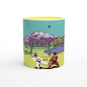 Può includere: Tazza in ceramica con interno giallo. L'esterno presenta una scena di arti marziali pixelata con due figure in un dojo. Lo sfondo include una montagna e un cancello.