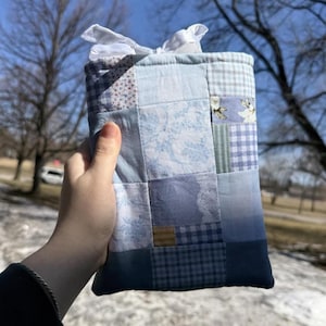 May include: A patchwork fabric tissue box cover in shades of blue, white, and a touch of gold. The cover is made of various patterned squares, including floral and gingham designs, and is tied at the top with a white ribbon.