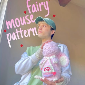 Peut inclure: Une peluche rose en forme de souris avec des détails blancs, une petite pochette et les mots "with love" brodés dessus. Le texte "Fairy mouse pattern" est écrit en rose au-dessus du jouet. Le jouet est tenu devant une personne portant une casquette verte.