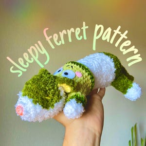 Könnte beinhalten: Ein handgehäkelter Frettchen in Grün und Weiß, mit dem Text "sleepy ferret pattern". Das Frettchen hat blaue Augen, rosa Wangen und eine rosa Nase. Der Körper besteht aus einer Mischung aus grünem und weißem Garn und wirkt weich und plüschig.