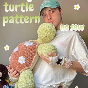 Könnte beinhalten: Eine handgefertigte Plüschschildkröte in Grün- und Brauntönen, verziert mit weißen Blumenakzenten. Der Text "fairy turtle pattern" und "no sew" sind sichtbar. Die Schildkröte wird von einer Person mit grüner Kappe gehalten.