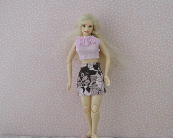 May include: A doll wearing a pink crop top with a ruffled neckline and a brown floral print mini skirt. The doll is also wearing pink sandals. The background is a pink dotted pattern.