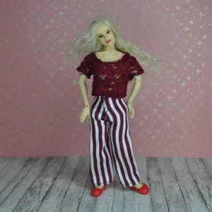 May include: A doll wearing a red crocheted top and white and red striped trousers. The doll has blonde hair and is standing on a wooden surface.
