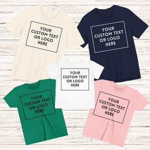 May include: Five short-sleeved t-shirts in cream, navy, white, green, and pink. Each shirt has a black square with the text "YOUR CUSTOM TEXT OR LOGO HERE" in the centre. The shirts are displayed on a light wood surface.