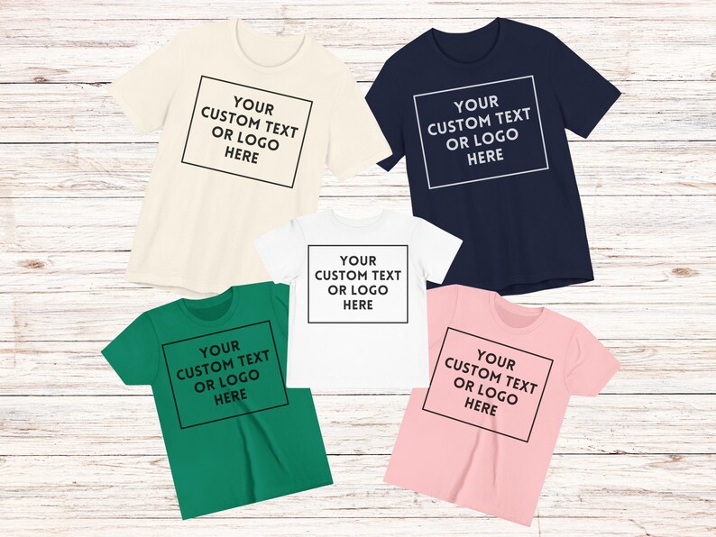 May include: Five short-sleeved t-shirts in cream, navy, white, green, and pink. Each shirt has a black square with the text "YOUR CUSTOM TEXT OR LOGO HERE" in the centre. The shirts are displayed on a light wood surface.