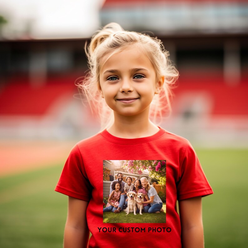 May include: A young girl wearing a red t-shirt featuring a family photo with two dogs. The text "YOUR CUSTOM PHOTO" is printed below the photo. The girl smiles, set against a blurred background of a sports field. The t-shirt is a vibrant red.
