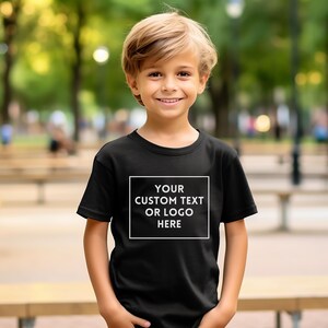 May include: A black t-shirt with a white rectangular design on the front. The design features the text "YOUR CUSTOM TEXT OR LOGO HERE" in a bold, sans-serif font. The t-shirt is short-sleeved and is worn by a young person.