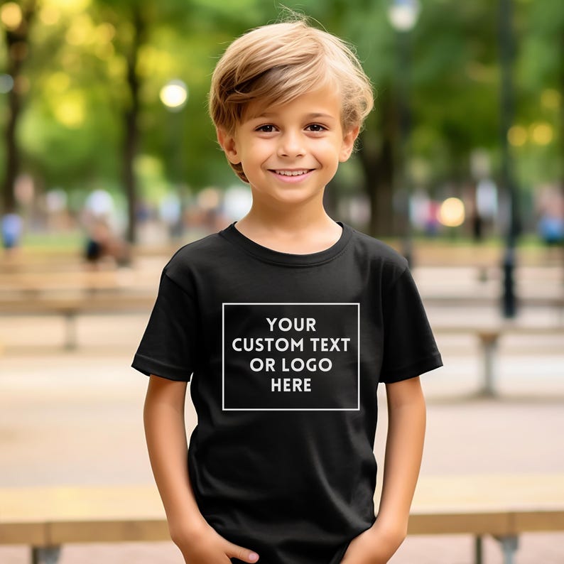 May include: A black t-shirt with a white rectangular design on the front. The design features the text "YOUR CUSTOM TEXT OR LOGO HERE" in a bold, sans-serif font. The t-shirt is short-sleeved and is worn by a young person.