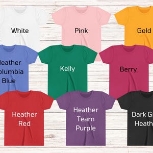 May include: A selection of short-sleeved t-shirts in a range of colours: white, pink, gold, blue, green, red, purple, and dark grey. Each shirt has a different colour name printed on it in a bold, sans-serif font. The shirts are displayed on a light wood-grain surface.