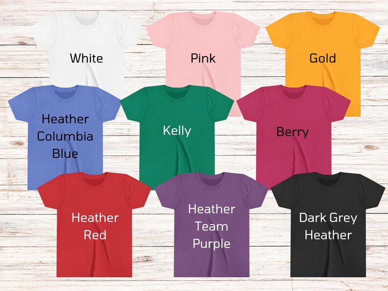 May include: A selection of short-sleeved t-shirts in a range of colours: white, pink, gold, blue, green, red, purple, and dark grey. Each shirt has a different colour name printed on it in a bold, sans-serif font. The shirts are displayed on a light wood-grain surface.
