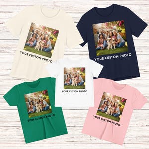 May include: Five short-sleeved t-shirts in cream, navy, white, green, and pink. Each shirt has a square image of a family with a dog, with the text "YOUR CUSTOM PHOTO" below. The shirts are on a light wood surface.