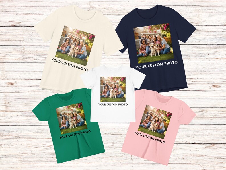 May include: Five short-sleeved t-shirts in cream, navy, white, green, and pink. Each shirt has a square image of a family with a dog, with the text "YOUR CUSTOM PHOTO" below. The shirts are on a light wood surface.