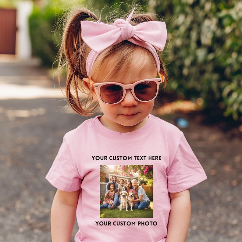 May include: A child's pink t-shirt featuring a family photograph and the text "YOUR CUSTOM TEXT HERE" and "YOUR CUSTOM PHOTO". The child is also wearing pink sunglasses and a pink bow headband.