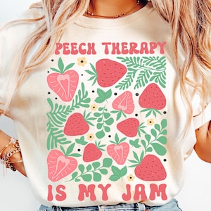 May include: Ivory-colored t-shirt with a graphic design featuring pink strawberries and green leaves. The text "SPEECH THERAPY IS MY JAM" is displayed in pink. The shirt is made of a soft material.