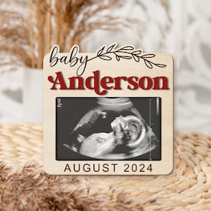 May include: A wooden photo frame with a white background and a black and white ultrasound image. The frame has the text "baby Anderson" in red and black lettering and "AUGUST 2024" in black lettering.