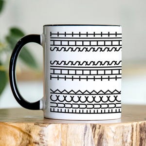 May include: White ceramic coffee mug with a black handle. The mug has a black and white geometric pattern of lines, waves, and zigzags.