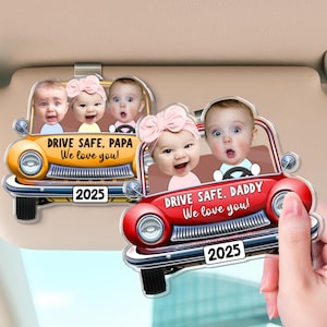 May include: Two car-shaped sun visor ornaments. One is yellow, the other red. Each features images of babies and the text "DRIVE SAFE, PAPA We love you!" and "DRIVE SAFE, DADDY We love you!" with the year 2025.