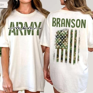 May include: Ivory-colored t-shirt with the words "ARMY mom" in green script and block letters on the front. The back of the shirt features the name "BRANSON" above a camouflage American flag design. The shirt has short sleeves and a relaxed fit.