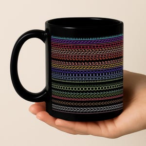 May include: Black ceramic mug featuring a colorful, patterned design of horizontal lines in various shapes and colors. The mug has a curved handle and is held by a hand.