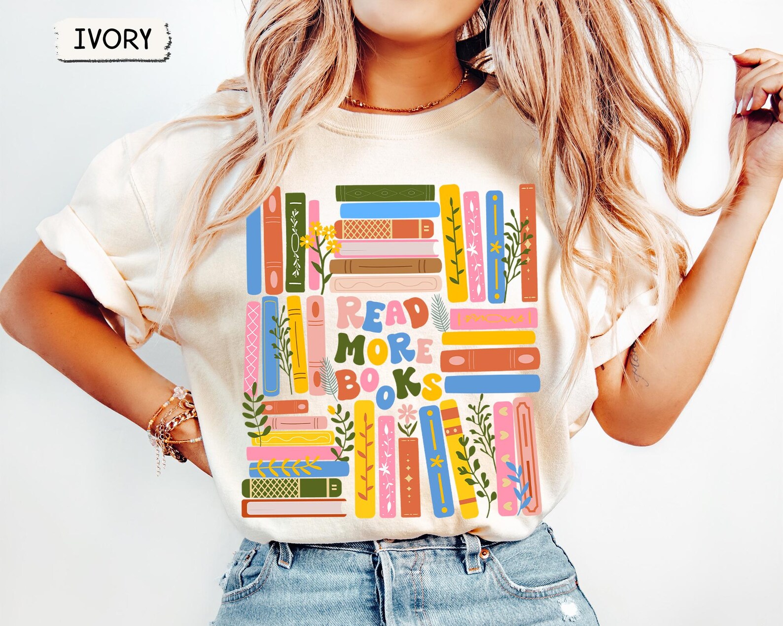 A woman wears an ivory-colored t-shirt that features a vibrant and whimsical design of books arranged in shelves. Each book spine is brightly colored with patterns, florals, stars, or vines, creating a playful and eye-catching display. In the center of the design, large multicolored letters spell out “READ MORE BOOKS.” The model has long, wavy blonde hair, rolled sleeves, light-wash jeans, and gold bracelets on her wrist. The overall aesthetic is cheerful and bookish, perfect for reading lovers and teachers.