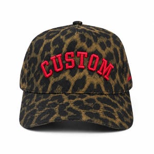 May include: A baseball cap featuring a brown and black leopard print pattern across the entire surface. The front panel displays the word CUSTOM embroidered in bold, raised red capital letters with a structured, arched design and a curved brim.