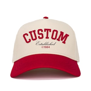 Puede incluir: Gorra de béisbol color crema con visera roja. La palabra "CUSTOM" está impresa en letras rojas en la parte delantera, con "Established ©1984" en letra más pequeña debajo. La gorra tiene una visera curva y un botón en la parte superior.