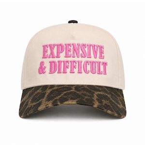 Peut inclure: Une casquette de baseball couleur crème avec le texte brodé "EXPENSIVE & DIFFICULT" en fil rose vif. La casquette présente un motif imprimé léopard marron et noir sur la visière incurvée et un petit bouton foncé au sommet.