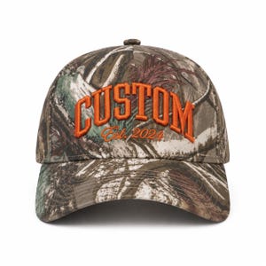 Peut inclure: Une casquette de baseball avec un motif camouflage dans des tons de marron, vert et blanc. L'avant présente le mot CUSTOM en broderie orange épaisse et en relief, avec le texte Est. 2024 brodé en orange dans une police cursive plus petite.